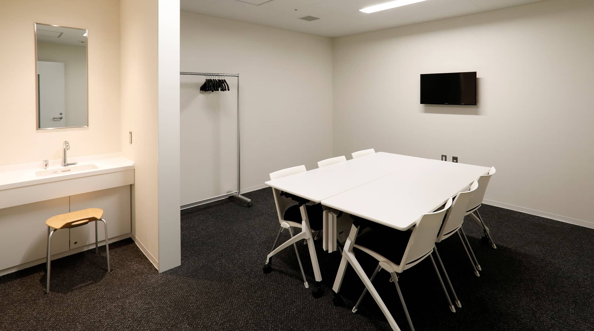 控室。壁側に洗面台とハンガーがあり、中央に白いテーブルと椅子6脚が配置され、テーブル横の壁にモニターが設置されている様子