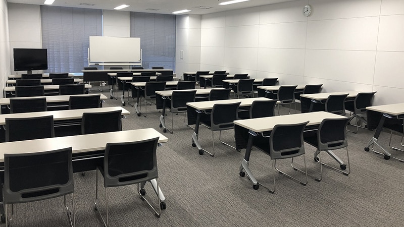 机と椅子がスクール形式に配置された会議室。正面にホワイトボードとモニターを備え、窓にはブラインドが設置されている。