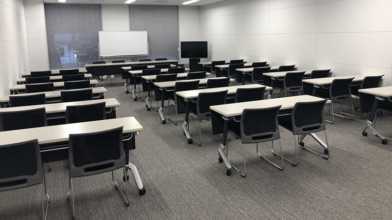 机と椅子がスクール形式に配置された会議室。正面にホワイトボードとモニターを備え、窓にはブラインドが設置されている。